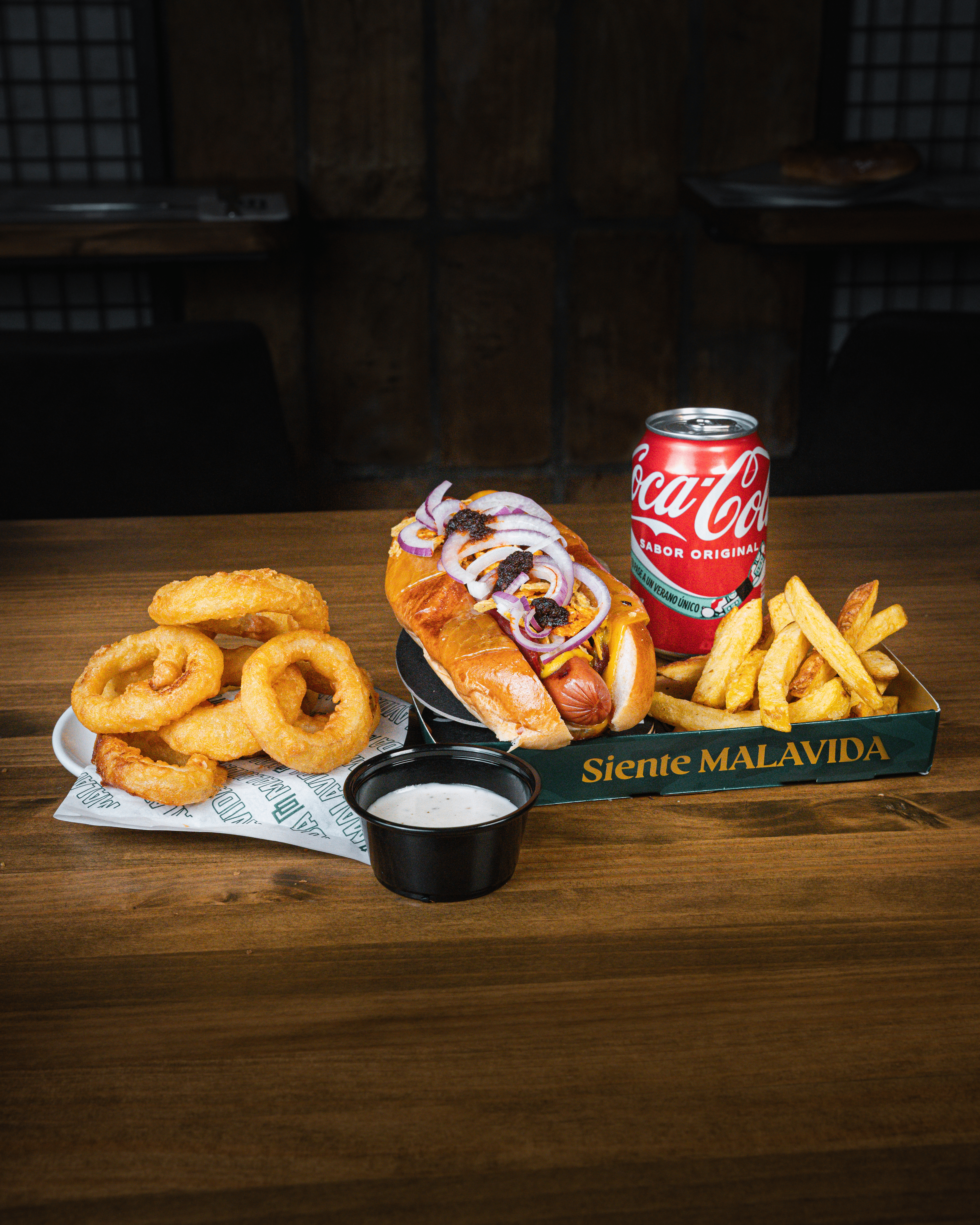 Imagen de un combo de comida rápida que incluye un perrito caliente con aderezos, papas fritas y una lata de Coca-Cola, presentado en una bandeja de Malavida.