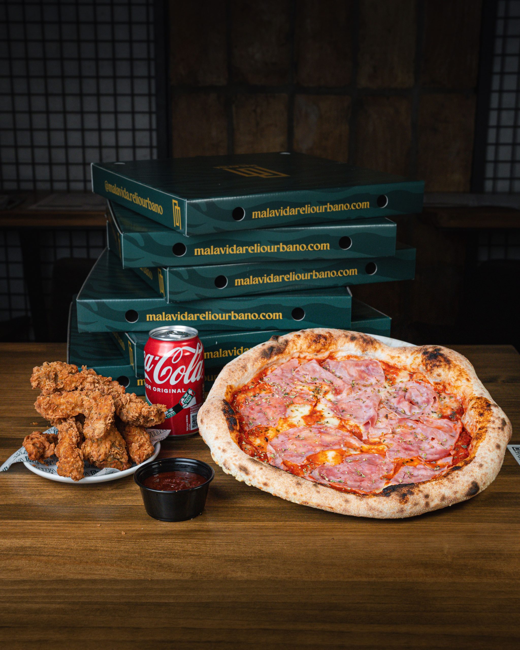 Imagen de un combo de comida que incluye una pizza de jamón y queso, crujientes tiras de pollo, una salsa y una lata de Coca-Cola, junto a varias cajas verdes apiladas de Malavida.